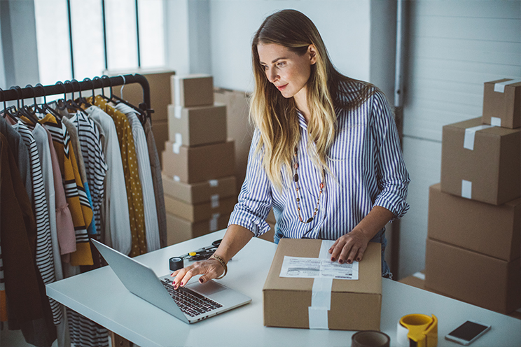 woman is surrounded by boxes and clothing as she stands to work at a computer